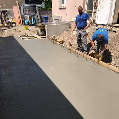 Zwei Arbeiter in blauen Hemden bauen an einem Baustellenbereich mit Stahlstangen und frisch gegossenem Beton.